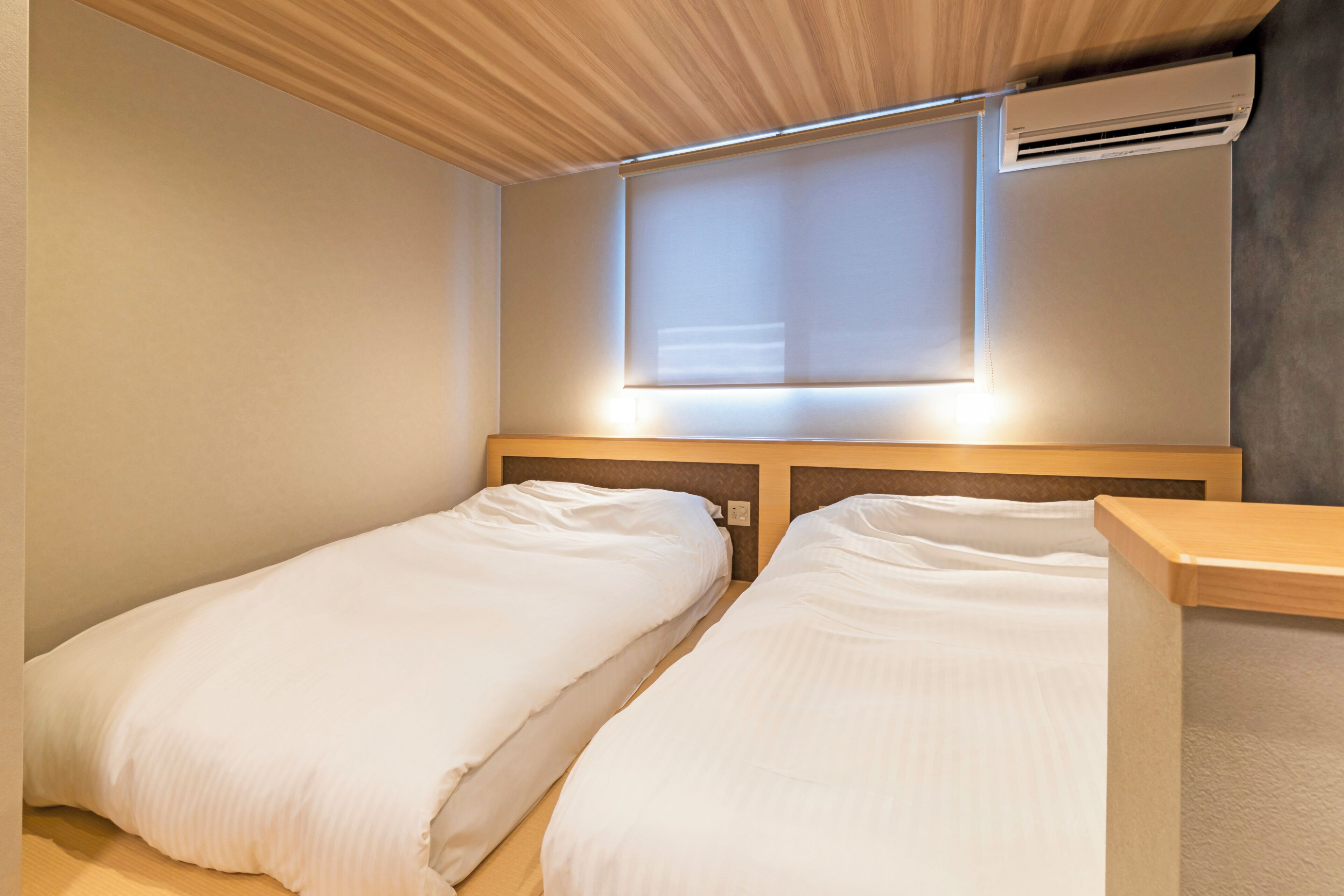 A room with two single beds, a wooden headboard, a window with blinds, and an air conditioning unit.