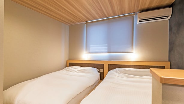 A room with two single beds, a wooden headboard, a window with blinds, and an air conditioning unit.
