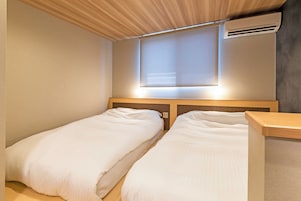A room with two single beds, a wooden headboard, a window with blinds, and an air conditioning unit.