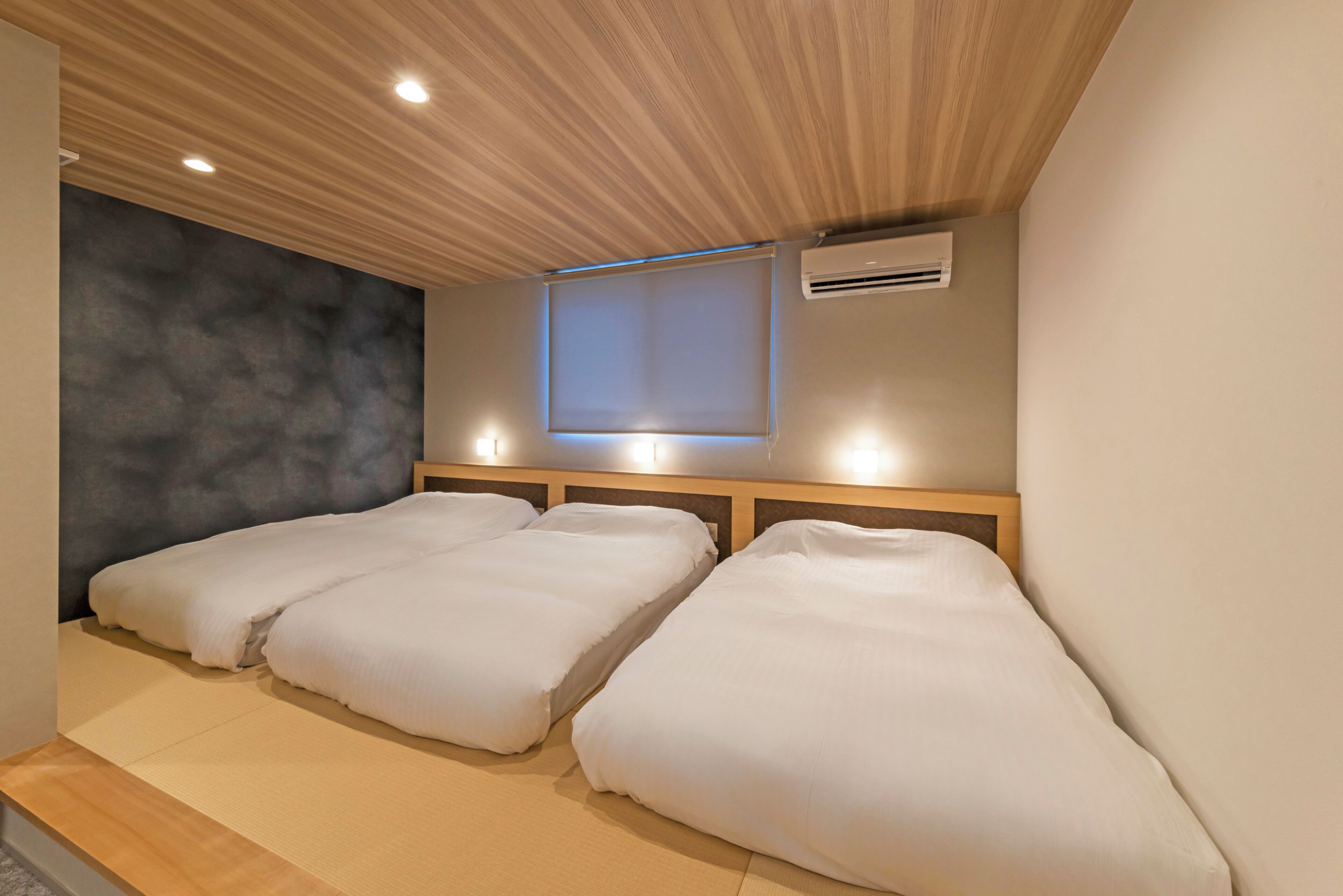 A room with three single beds, a wooden ceiling, an air conditioning unit, and a window with blinds.