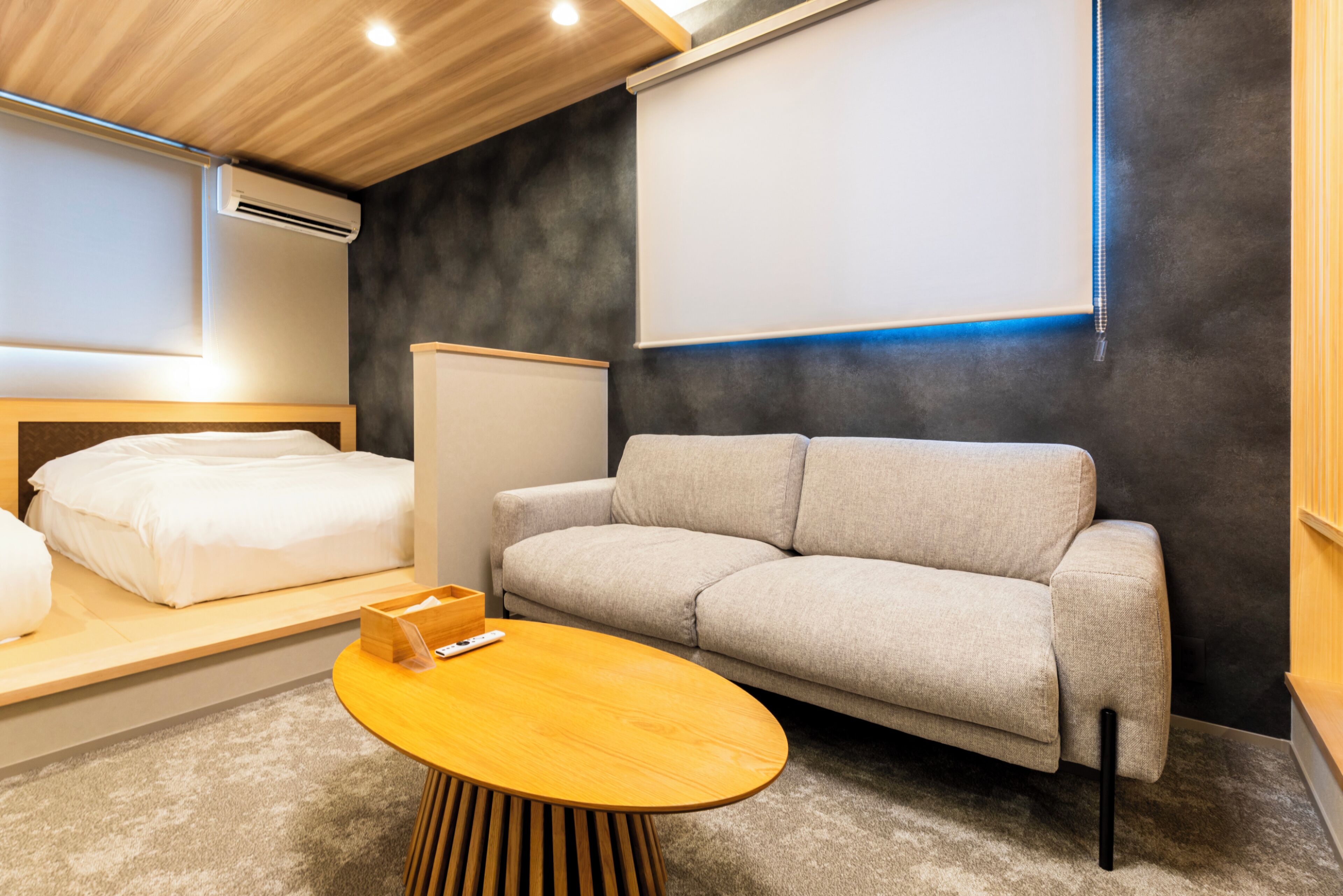 A modern hotel room with a sofa, a wooden coffee table, a bed, and a wall-mounted air conditioner.