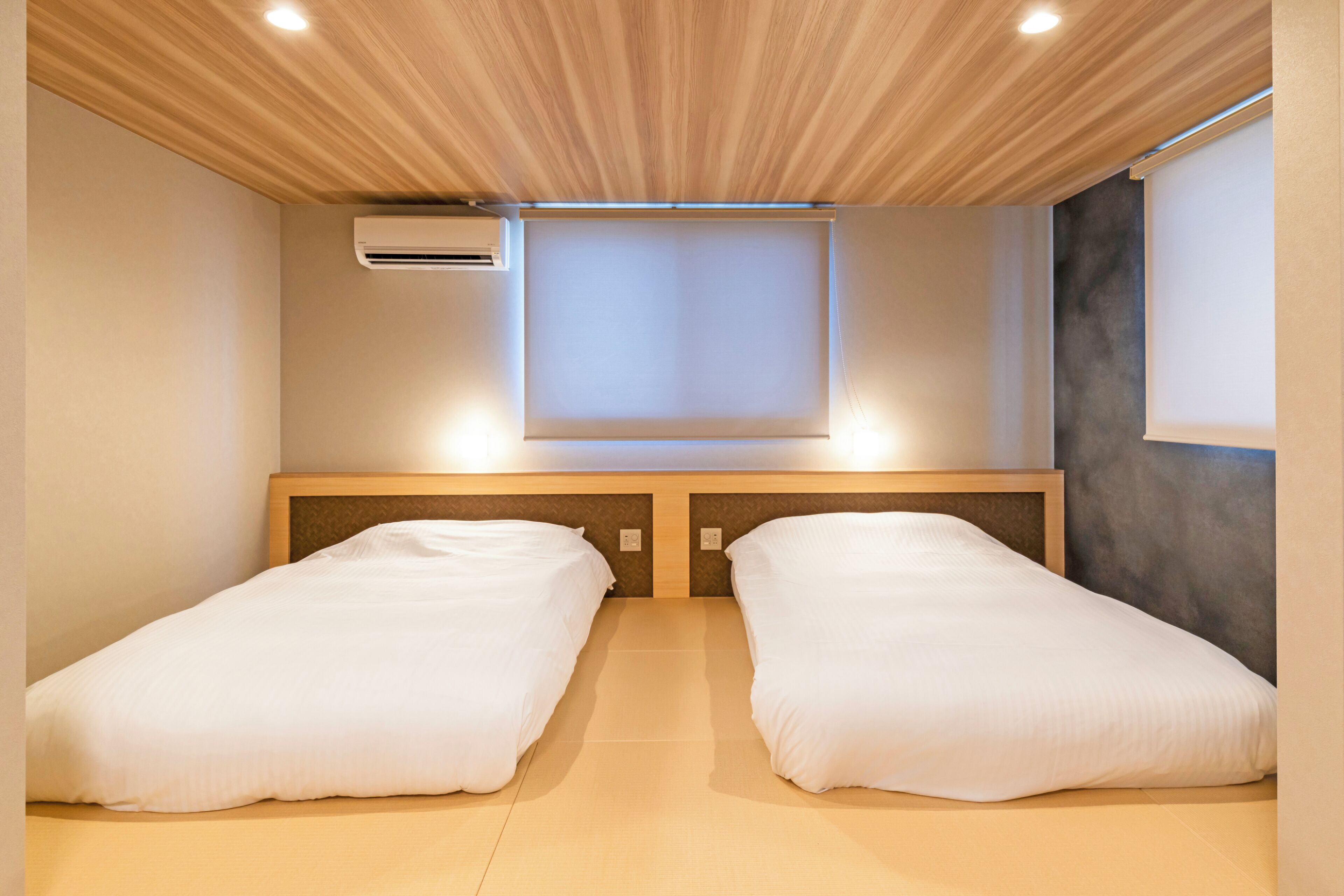 A room with two single beds, a wooden ceiling, an air conditioning unit, and a window.