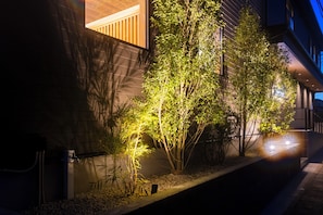 Vista nocturna de un edificio con jardín ajardinado, plantas de bambú y un elemento acuático.