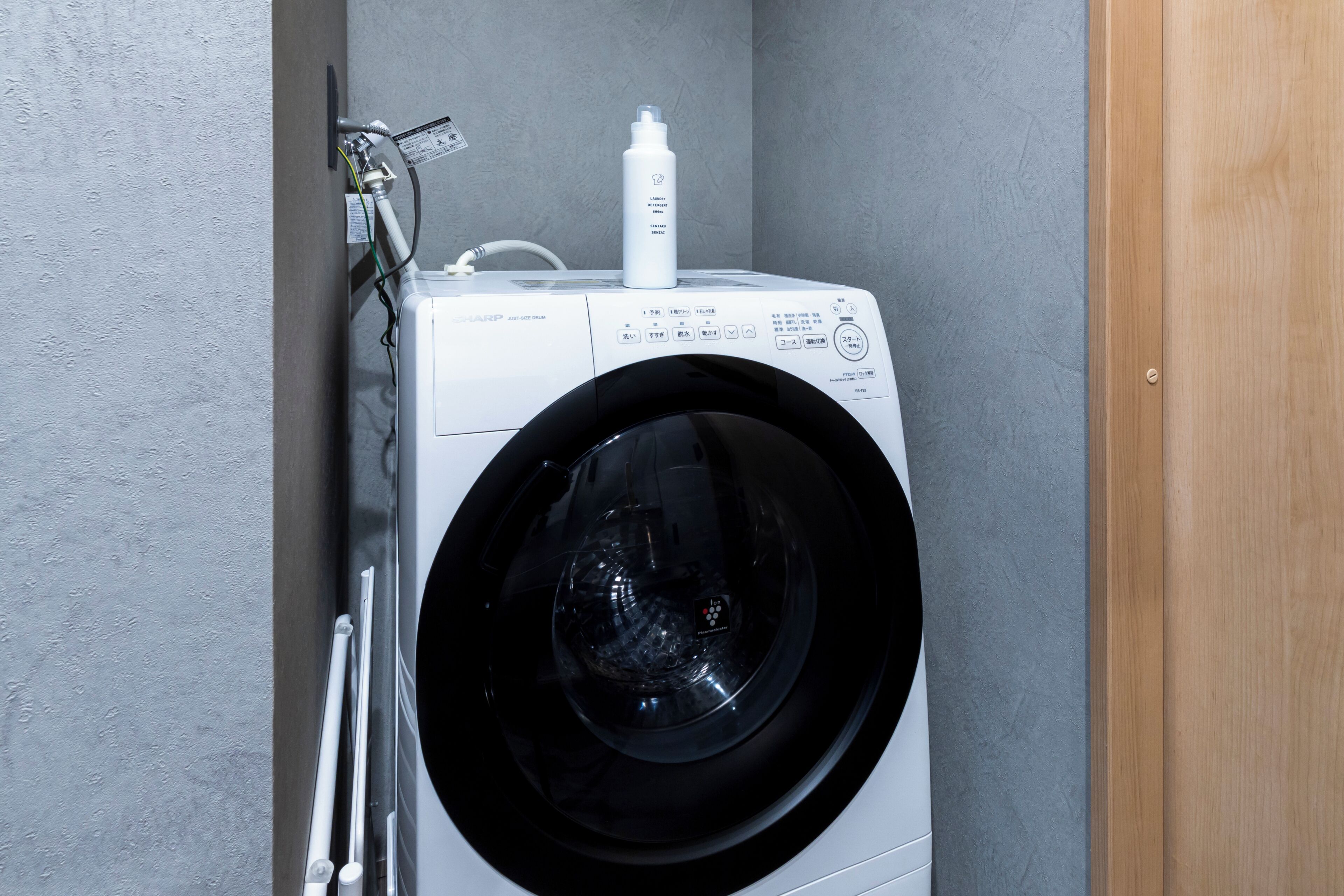 A front-loading washing machine with a control panel, a detergent bottle, and a hose connected to a wall-mounted electrical outlet.