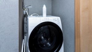 A front-loading washing machine with a control panel, a detergent bottle, and a hose connected to a wall-mounted electrical outlet.
