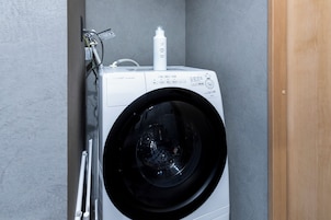 A front-loading washing machine with a control panel, a detergent bottle, and a hose connected to a wall-mounted electrical outlet.