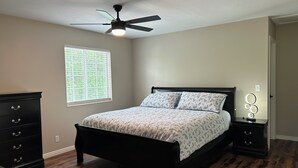 Ein Schlafzimmer mit einem Bett, einem Deckenventilator, einem Nachttisch mit Lampe, einem Schminktisch und einem Fenster mit Jalousien.