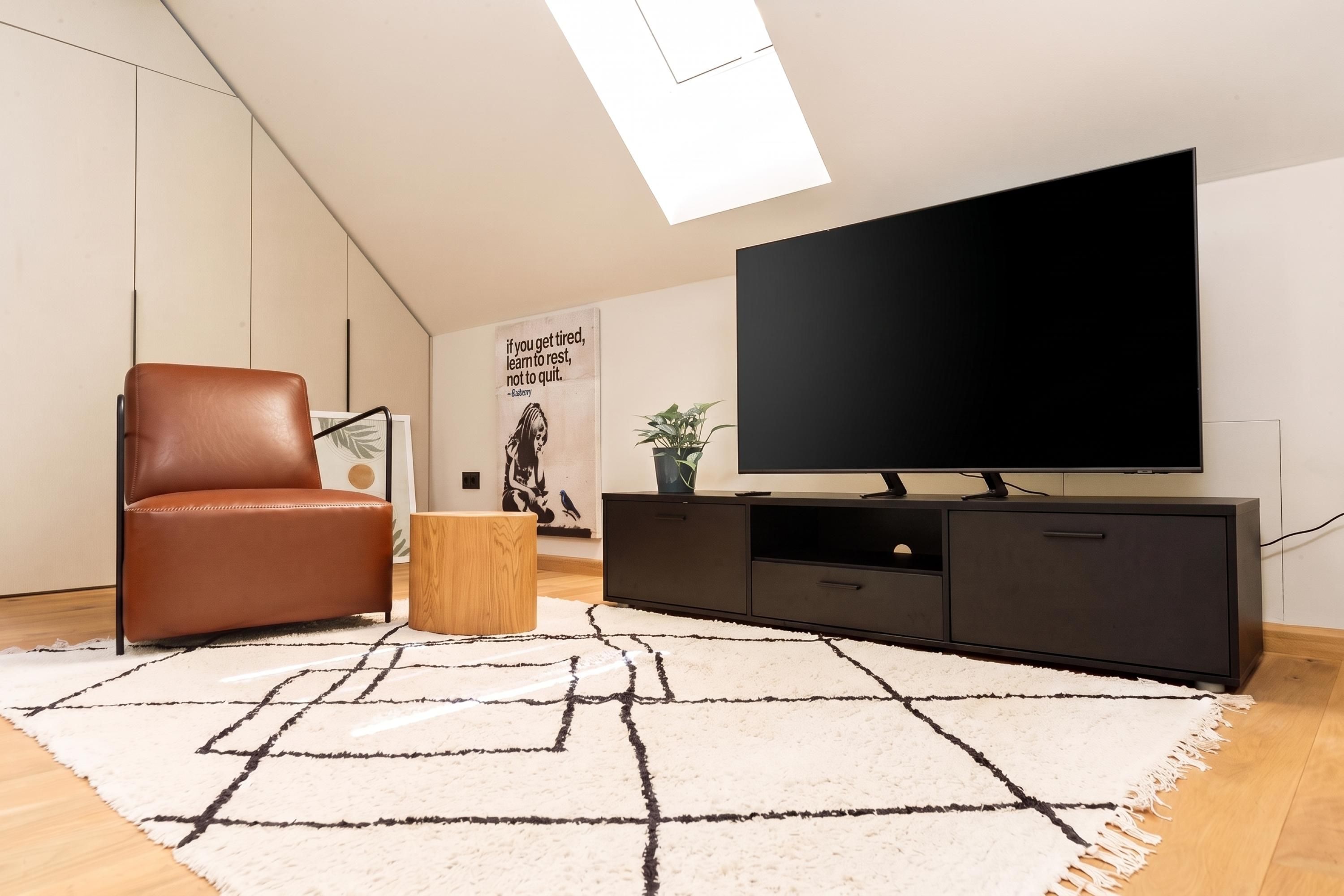 Una sala moderna con un sillón de cuero, un televisor de pantalla plana sobre un mueble bajo y una alfombra de diseño geométrico.