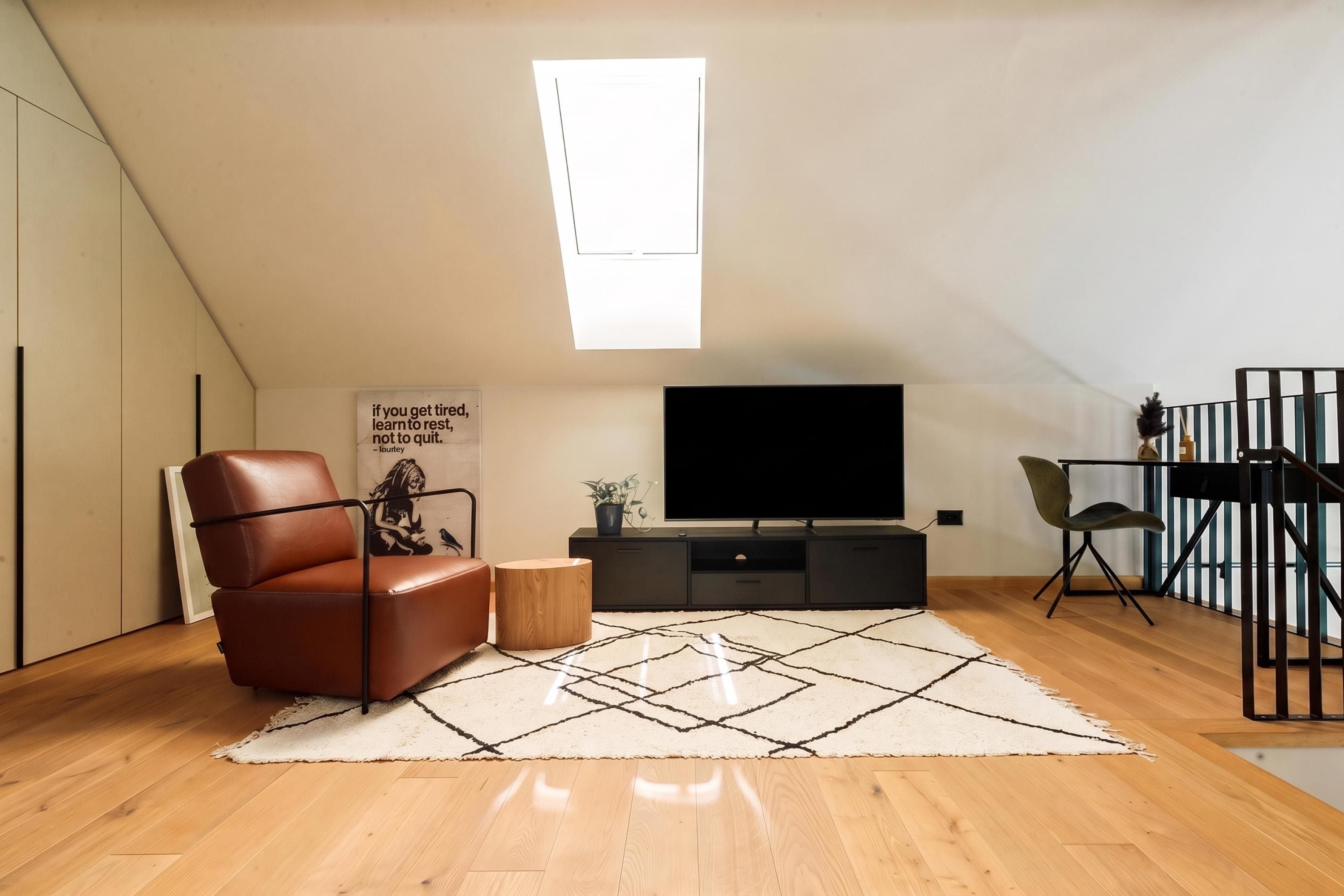 Ein modernes Wohnzimmer mit schrägem Dach, einem großen Flachbildfernseher, einem Ledersessel, einem kleinen Holztisch und einem Teppich mit geometrischem Muster.