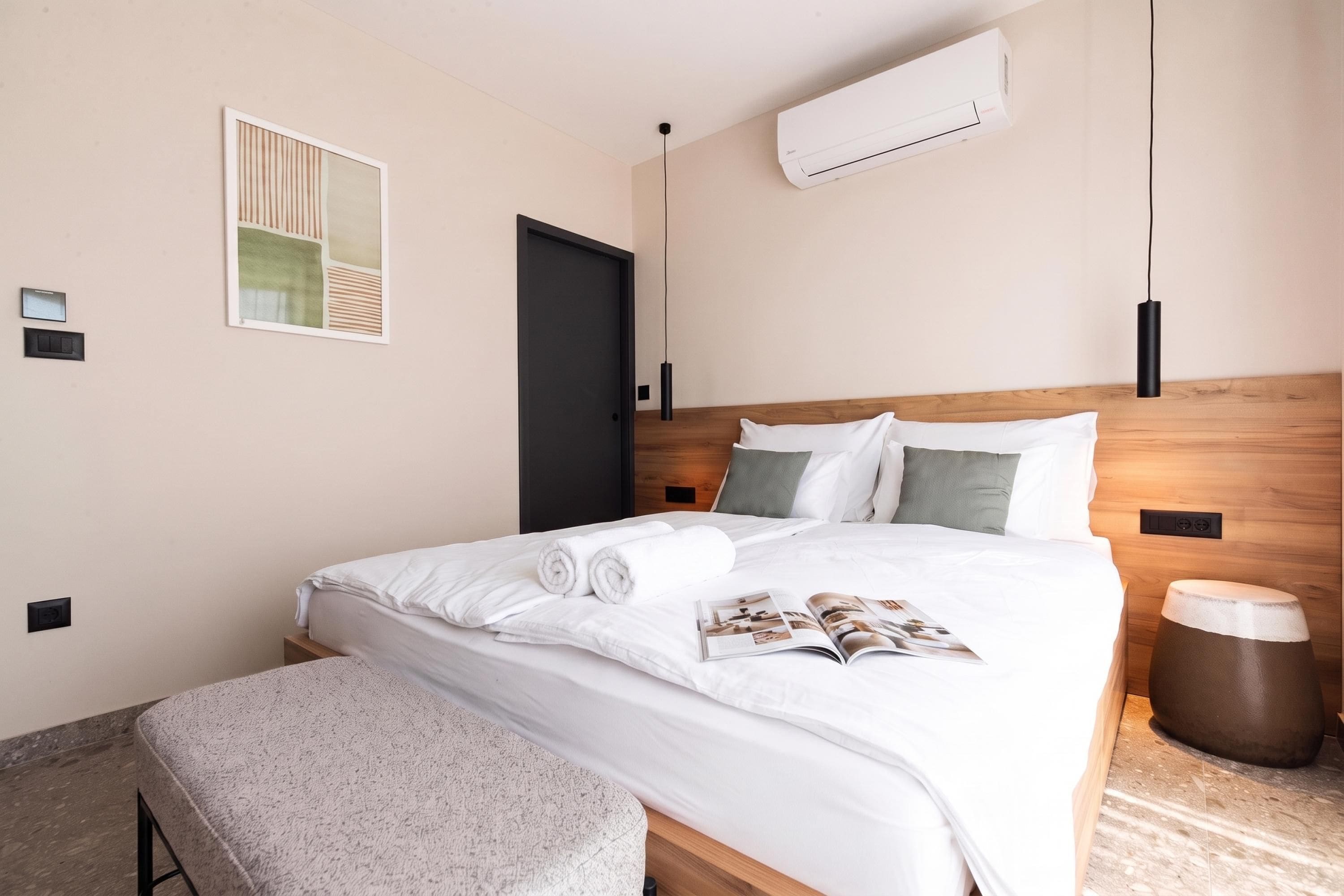 A neatly made bed with white linens and a magazine, a wooden headboard, a wall-mounted air conditioner, and a small bedside table with a lamp.