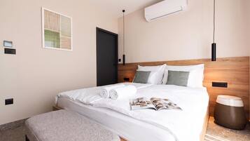 A neatly made bed with white linens and a magazine, a wooden headboard, a wall-mounted air conditioner, and a small bedside table with a lamp.