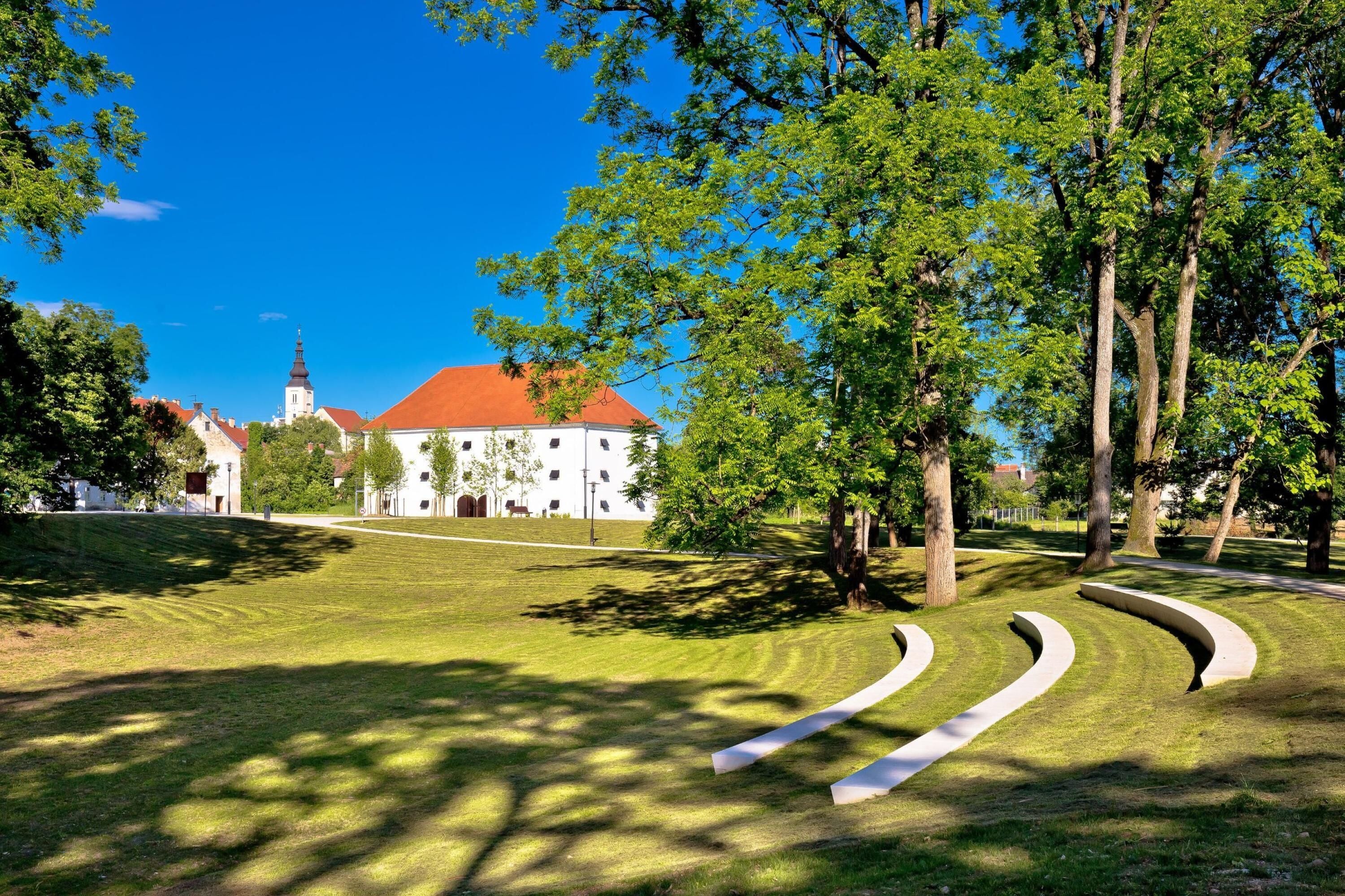 A park with a white building with a red roof, surrounded by trees and grass.