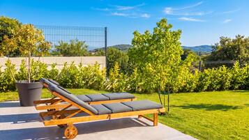 A sun lounger with wheels on a patio, surrounded by greenery and a clear sky.