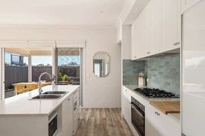 A modern kitchen with a built-in stove, a sink, and a dining area with a view. - Palm Cove—Tranquil Coastal Retreat with Fire Pit (Dunsborough)