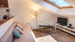 A compact living space with a sloped ceiling, a sofa with cushions, a standing lamp, a glass table, a flat-screen TV, and a wooden floor. - Ema Studio on the Main Promenade (Koper)