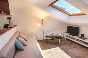 Ein kompakter Wohnbereich mit schrägverdeckter Decke, einer Couch mit Kissen, einer Stehlampe, einem Glastisch, einem Flachbildfernseher und einem Holzboden.