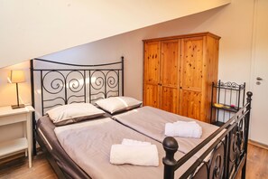 Ein Doppelbett mit Metallgestell, auf jedem Bett zwei Kissen und ein gefaltetes Handtuch. Ein Holzschrank und ein kleines Regal mit einer Lampe und einer Haarbürste sind ebenfalls vorhanden.