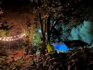 Escena nocturna al aire libre con piscina, luces colgantes y una estructura de techo de paja.