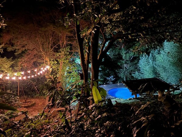 Escena nocturna al aire libre con piscina, luces colgantes y una estructura de techo de paja.