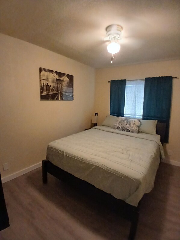A bedroom with a bed, a ceiling fan, a window with curtains, and a wall-mounted painting.