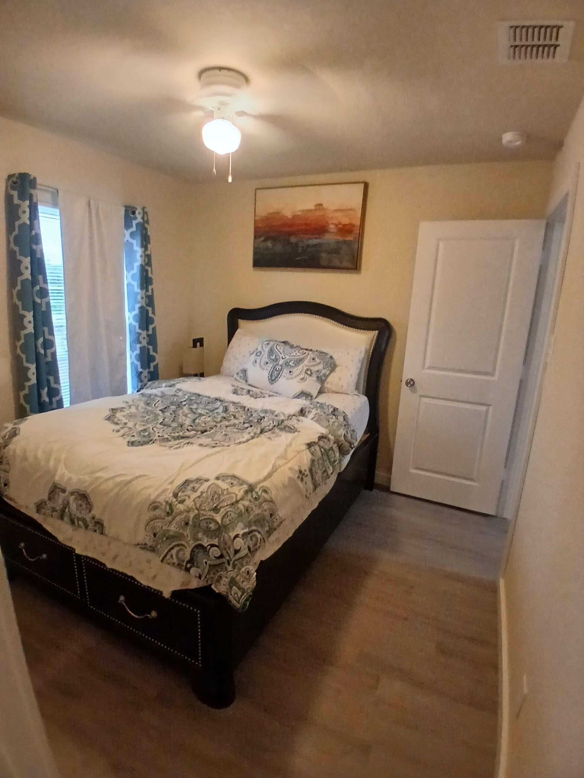 Quarto com cama, ventilador de teto, porta e janela com cortinas.