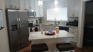 Una cocina moderna con un refrigerador de acero inoxidable, un microondas y una cafetera. Hay dos taburetes de bar y una pequeña isla con una cesta.