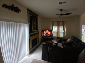 A living room with a fireplace, a television, a ceiling fan, and a window with curtains.