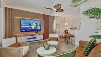 A modern living room with a flat-screen TV, comfortable seating, a dining area, and a kitchen in the background.