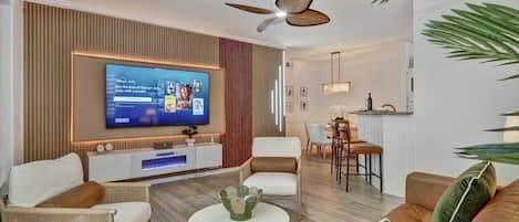 A modern living room with a flat-screen TV, comfortable seating, a dining area, and a kitchen in the background.