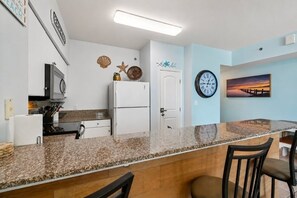 Una cucina con piano di lavoro in granito, mobili bianchi, un frigorifero e una sveglia appesa al muro.