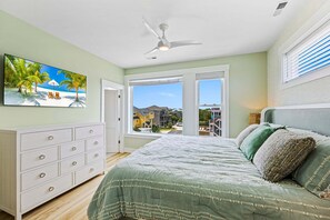 A bedroom with a bed, a dresser, a ceiling fan, and a window with a view of a residential area. - Coastal Bliss - Oceanfront w/ views, pool, hot tub, and easy beach access. (Corolla)
