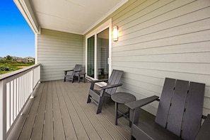 A wooden deck with two Adirondack chairs and a table, a glass door leading inside, and a light fixture on the wall. - Coastal Bliss - Oceanfront w/ views, pool, hot tub, and easy beach access. (Corolla)