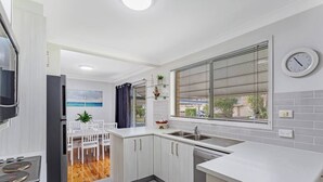 A modern kitchen with white cabinetry, a built-in dishwasher, a window with blinds, and a dining area with a white table and chairs. - Barefoot Bungalow Walk to Little Beach (Nelson Bay)