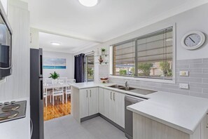 A modern kitchen with white cabinetry, a built-in dishwasher, a window with blinds, and a dining area with a white table and chairs. - Barefoot Bungalow Walk to Little Beach (Nelson Bay)