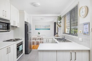 A modern kitchen with white cabinetry, stainless steel appliances, a dining area with a white table and chairs, and a window with blinds.
