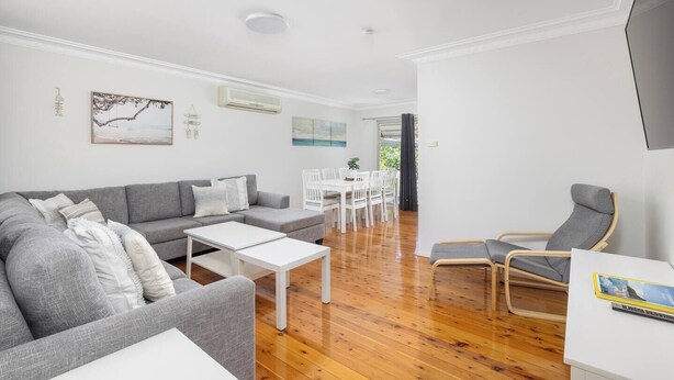A modern living room with a grey sofa, white coffee table, and a dining area with white chairs. - Barefoot Bungalow Walk to Little Beach (Nelson Bay)