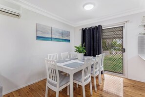 A dining area with a white table, chairs, and a potted plant. - Barefoot Bungalow Walk to Little Beach (Nelson Bay)