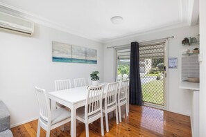 A dining area with a white table and chairs, a painting on the wall, and a view through a glass door.