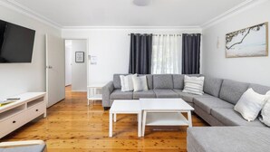 A modern living room with a grey sofa, white coffee table, and a flat-screen TV mounted on the wall. - Barefoot Bungalow Walk to Little Beach (Nelson Bay)