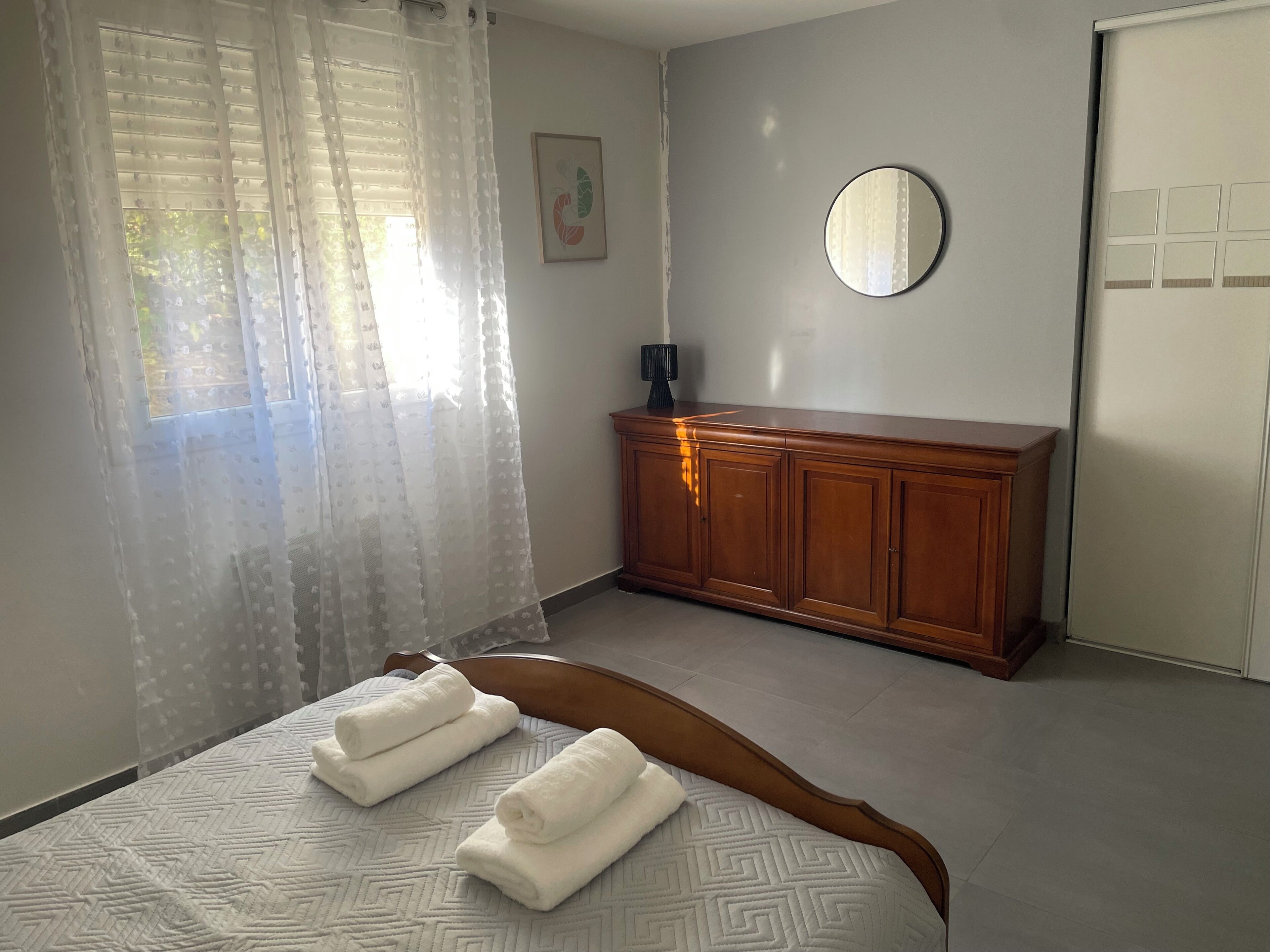 Un dormitorio con cama, cómoda de madera, espejo y ventana con cortinas translúcidas.