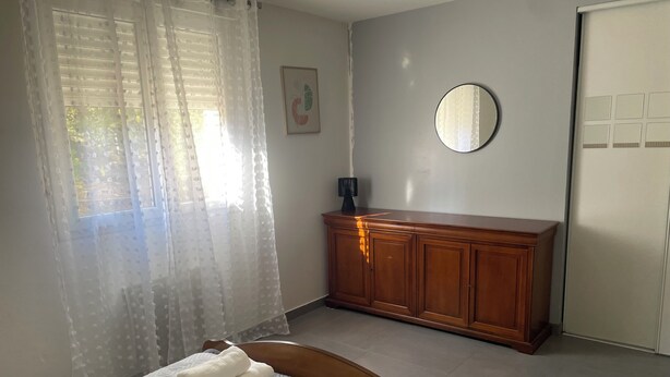 A bedroom with a bed, a wooden dresser, a mirror, and a window with sheer curtains. - 3-room NE apartment on the garden level (Varetz)