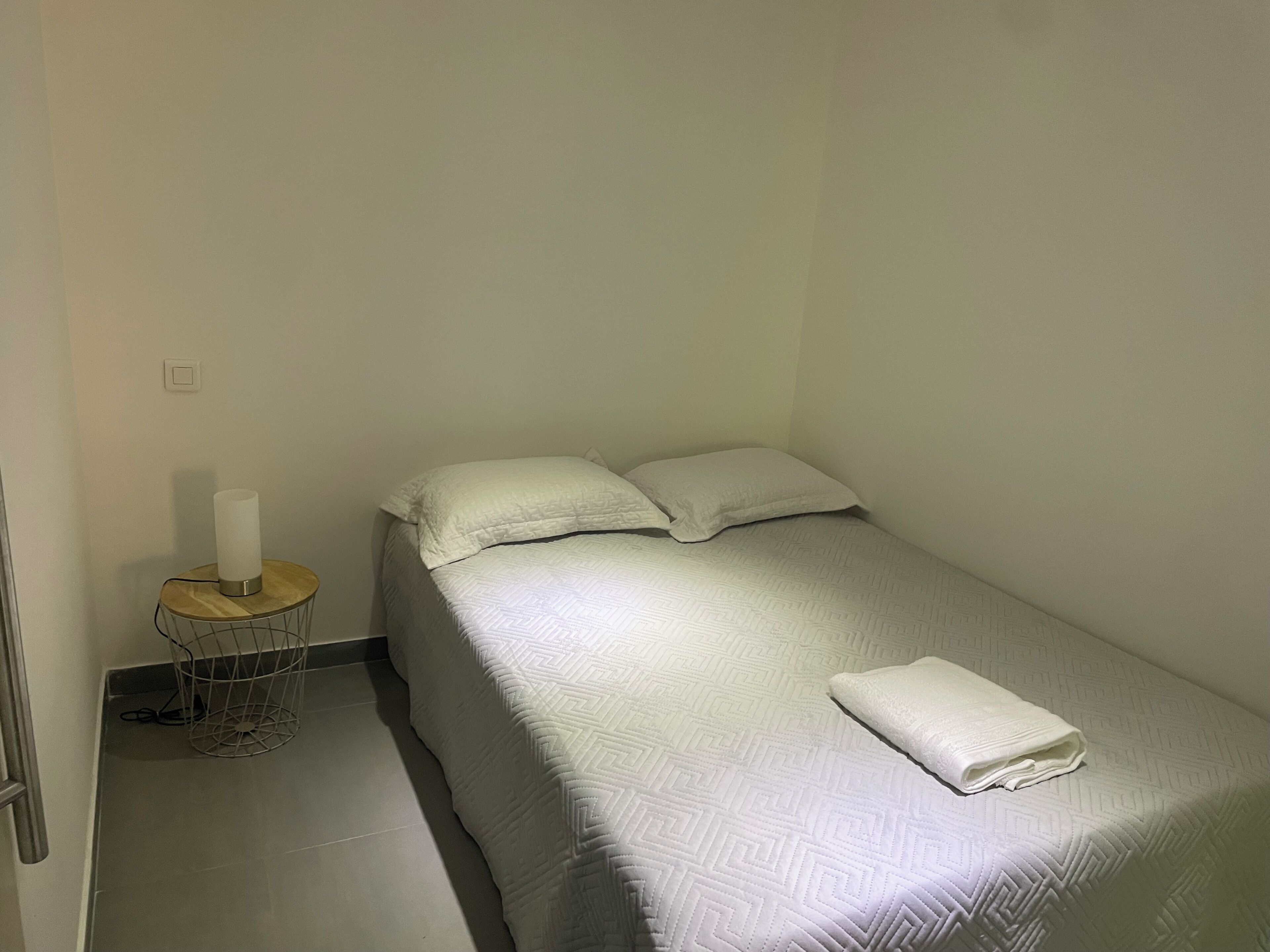 Una cama individual con colcha blanca, dos almohadas, una mesita de noche con lámpara y una toalla doblada sobre la cama.