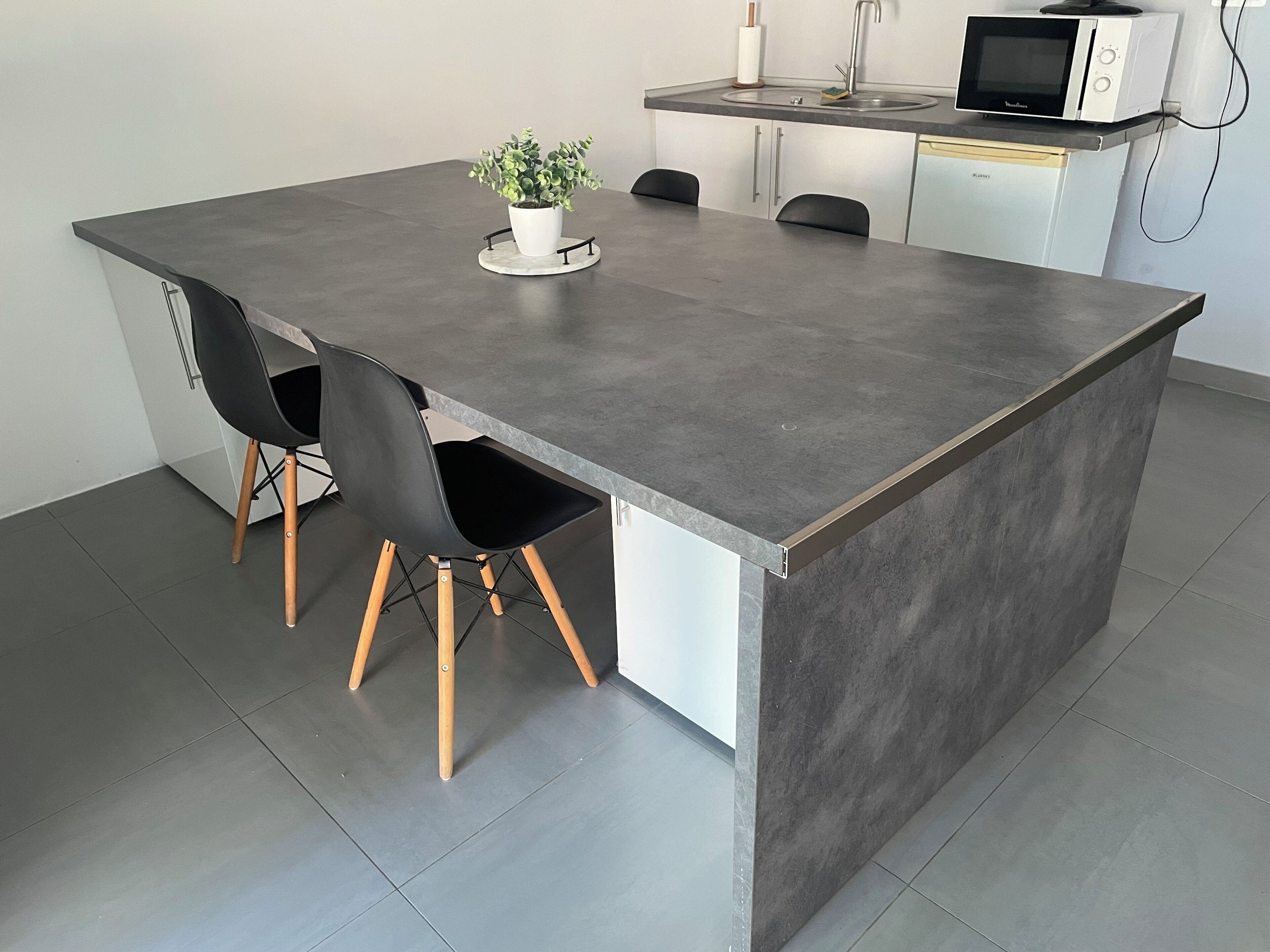Una cocina moderna con encimera gris, dos sillas negras, una planta en maceta y un microondas.