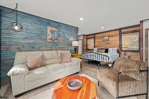 A living room with a sofa, armchair, and a coffee table. There is a bed with a white bedspread and a Texas flag. A painting is on the wall, and a lamp is on a side table. - Luckenbach Guesthouse, Hill Country Views! (Fredericksburg)