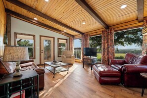 A spacious living room with a wooden ceiling, leather sofas, a television, and a large window offering a view of the outdoors. - Luckenbach House, Hot tub, Views! (Fredericksburg)