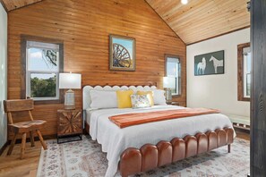 A bedroom with a wooden ceiling, a bed with a white headboard, two bedside lamps, and a window with a view of trees. - Luckenbach House, Hot tub, Fire pit, Views! (Fredericksburg)
