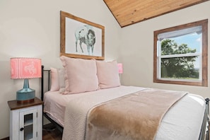 A bedroom with a bed, a nightstand, a lamp, a framed picture on the wall, and a window. - Luckenbach House, Hot tub, Views! (Fredericksburg)