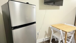 A compact kitchenette with a stainless steel refrigerator, a wooden dining table, and a wall-mounted television.