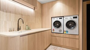 Pequeña lavandería con dos lavadoras de carga frontal, un lavabo con grifo y un estante con artículos de aseo.