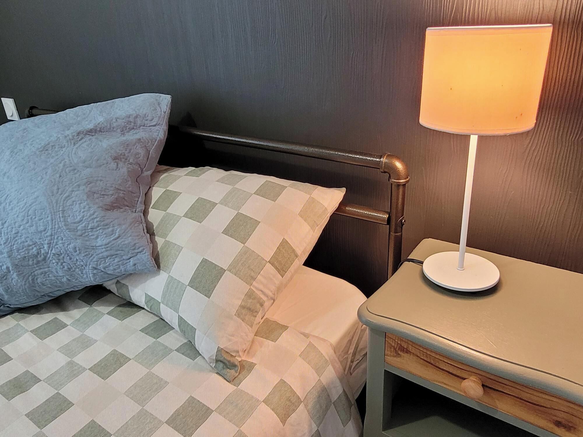 Ein Bett mit kariertem Überwurf und zwei Kissen, ein Nachttisch mit Lampe und ein Holznachtkästchen.